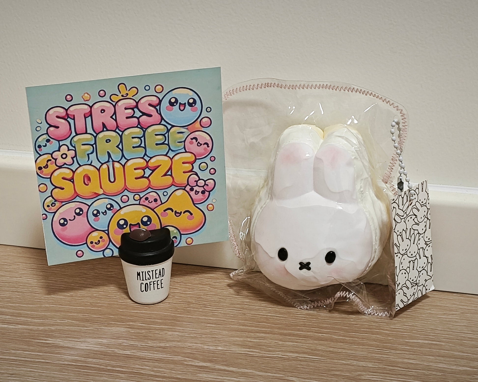 A white bunny-shaped squishy toy in clear plastic packaging sits on a wooden surface next to a colorful 'STRESS FREE SQUEEZE' card and a small black-and-white 'MILSTEAD COFFEE' cup. The background is a plain beige wall.