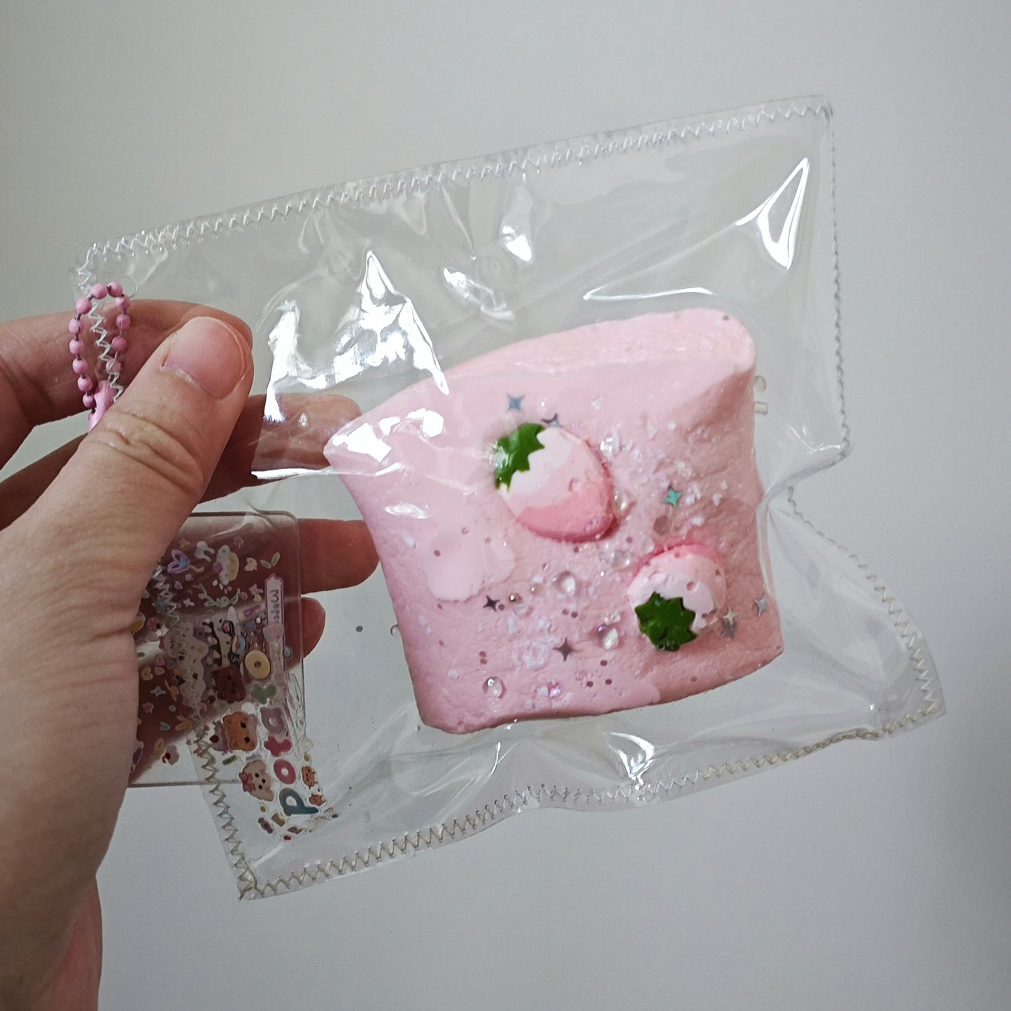 A hand holding a clear plastic bag containing a pink strawberry-themed squishy toy, with a small pink beaded charm and decorative glitter visible inside the bag against a plain light gray background.