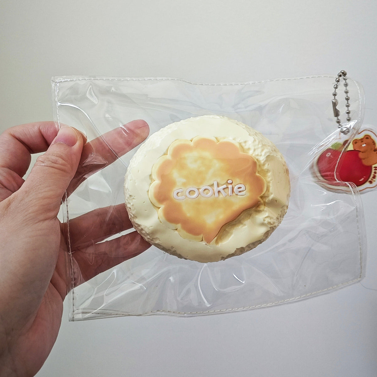 A hand holding a clear plastic bag containing a round, cream-colored cookie-shaped item with a golden-brown center and the word 'cookie' printed on it, alongside a small red fruit charm keychain.