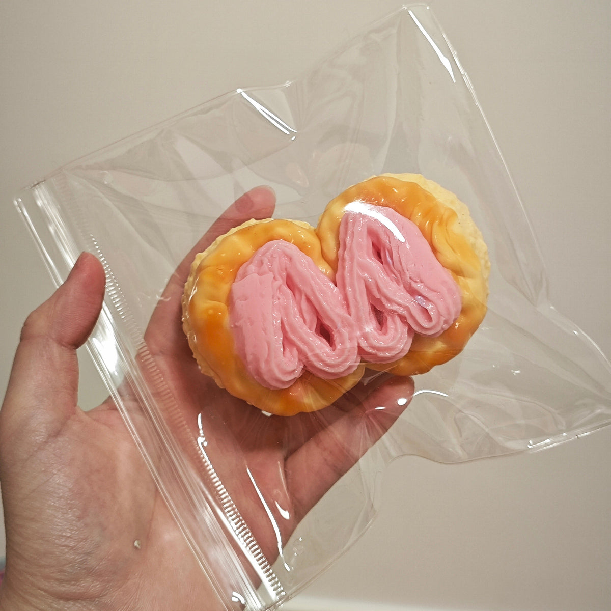 A hand holding a clear plastic bag containing a novelty squishy toy shaped like a pink-and-gold pastry, photographed against a plain light beige wall.