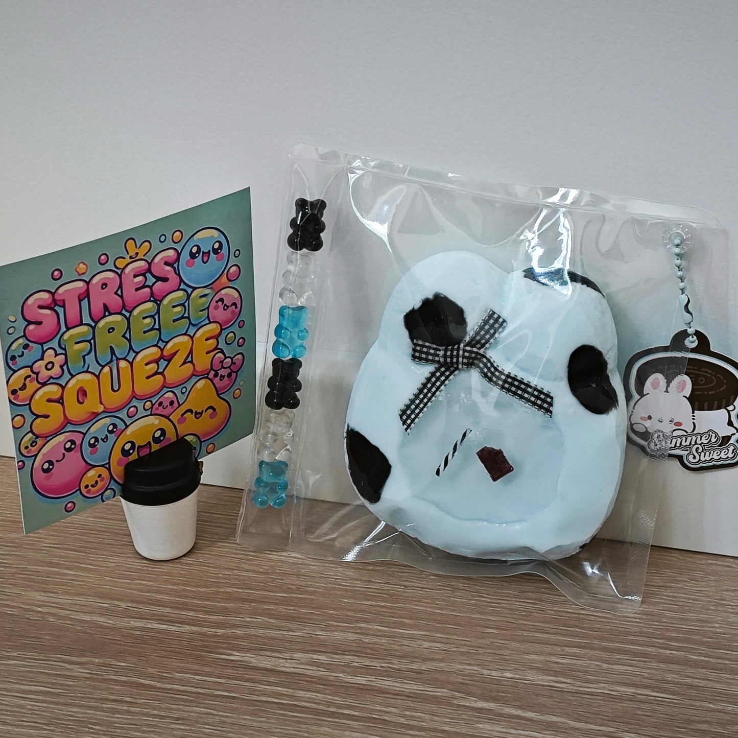 A light blue plush earbud case with black details and a black-and-white checkered bow, shown in a clear plastic bag with a keychain tag. A colorful 'STRESS FREE SQUEEZE' card, a small cup-shaped accessory, and a beaded bracelet are arranged beside it on a wood surface against a white wall.