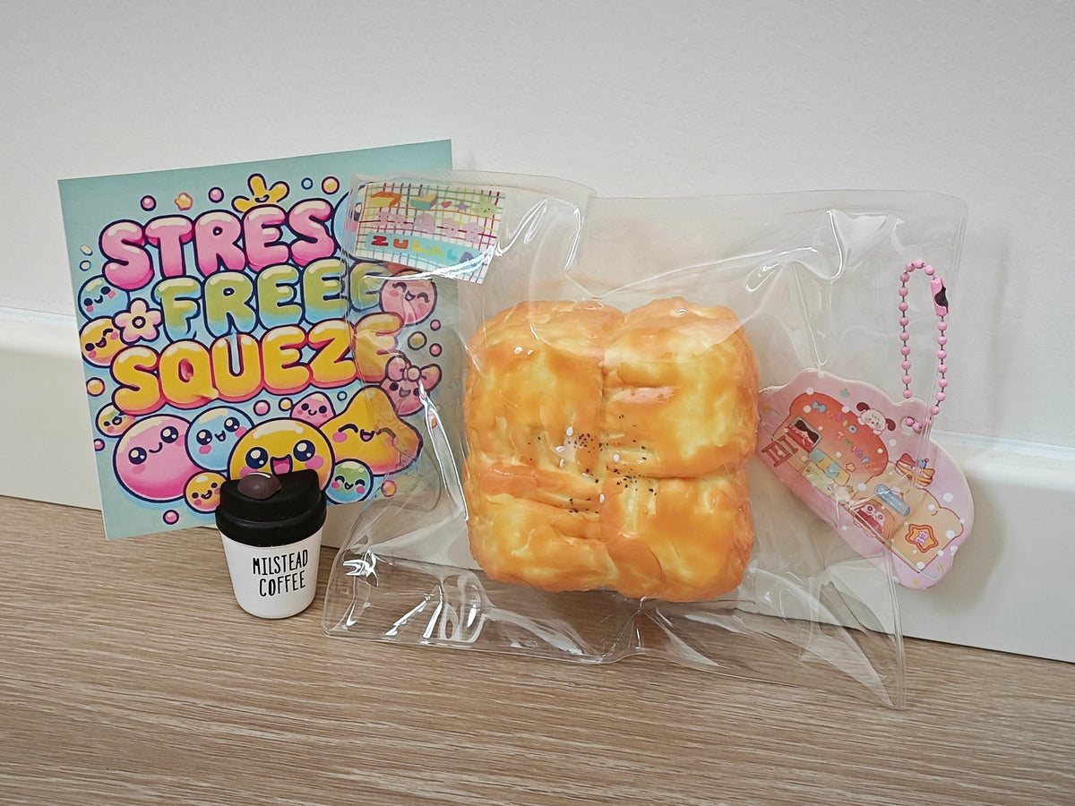 A Stres Free Squeeze toy set on a light wood surface against a white wall, featuring a yellow-orange bread-like squishy, a small coffee cup accessory labeled 'Missteam Coffee,' and a colorful printed card.
