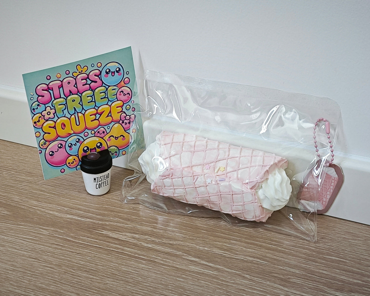 A pink and white quilted stress toy wrapped in clear plastic packaging, shown next to a small black coffee cup accessory and a colorful 'STRESS FREE SQUEEZE' card on a light wood floor against a white wall.