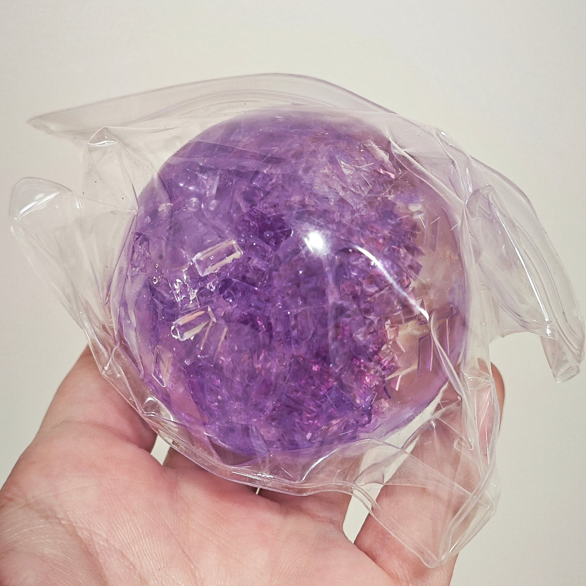 A hand holding a purple glittery crystal ball wrapped in clear plastic against a plain light background.