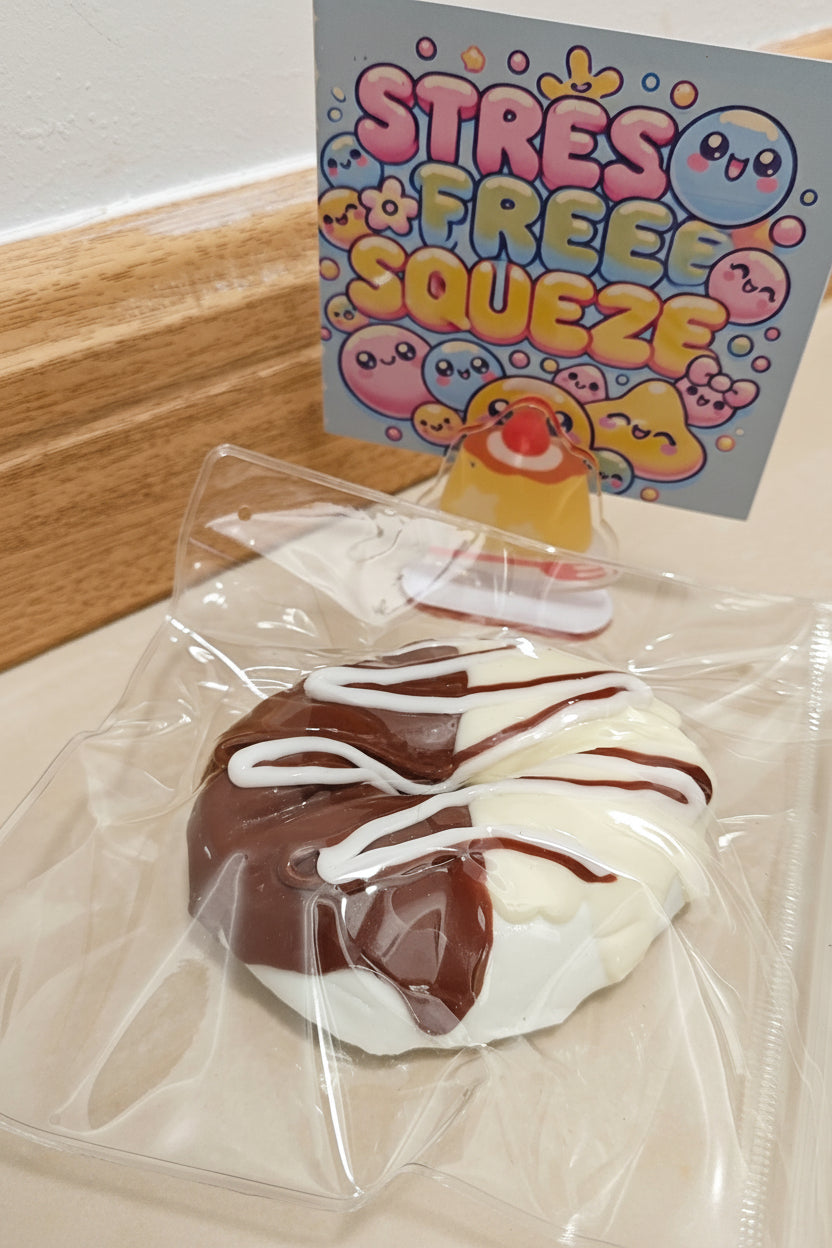 A packaged donut-shaped squishy toy with white and dark brown frosting-like sections and drizzled chocolate lines, sealed in a clear plastic bag. A small printed card behind it reads 'STRESS FREE SQUEEZE' with cartoon characters.