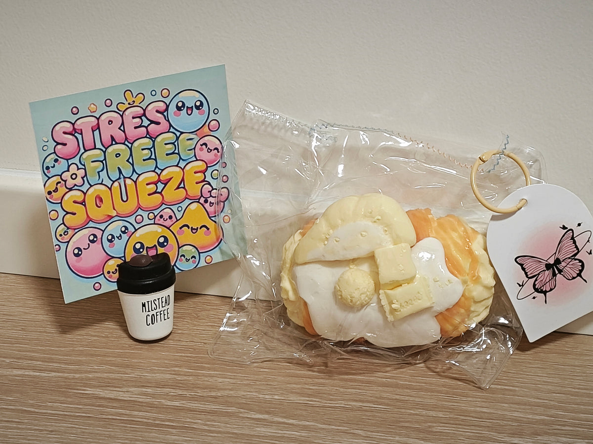 A cute pastel-colored squishy toy shaped like a pastry with butter and cream, shown in a clear plastic bag with a butterfly tag. Next to it is a small white cup labeled 'MISTYAR COFFEE' and a colorful card that reads 'STRESS FREE SQUEEZE'.