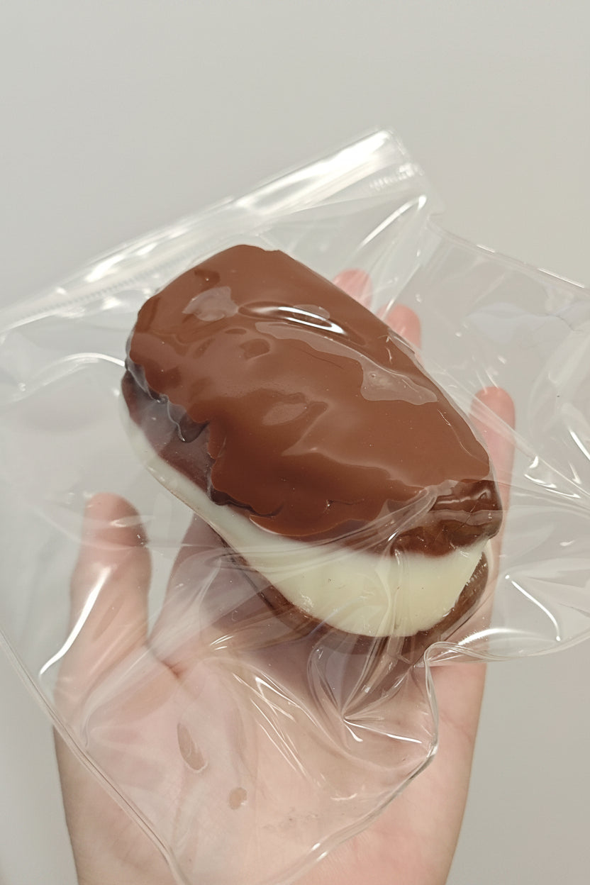 A hand holding a chocolate-covered ice cream sandwich inside a clear plastic bag against a plain light gray background.