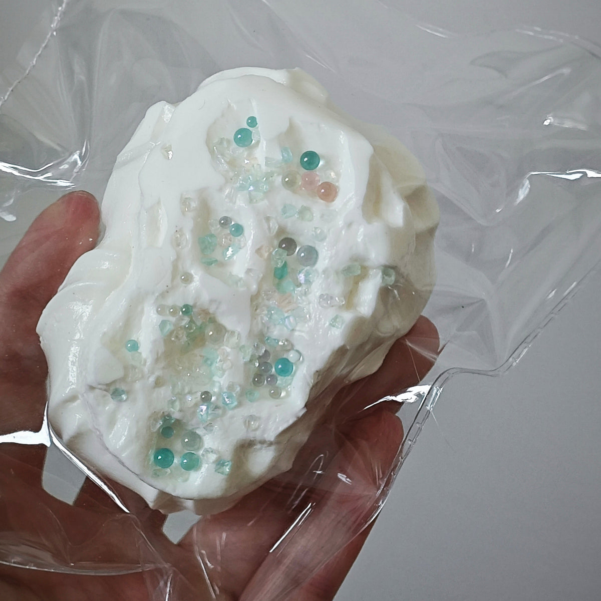 A hand holding a white, textured squishy toy or foam block decorated with small translucent teal and pink beads, wrapped in clear plastic against a plain light background.