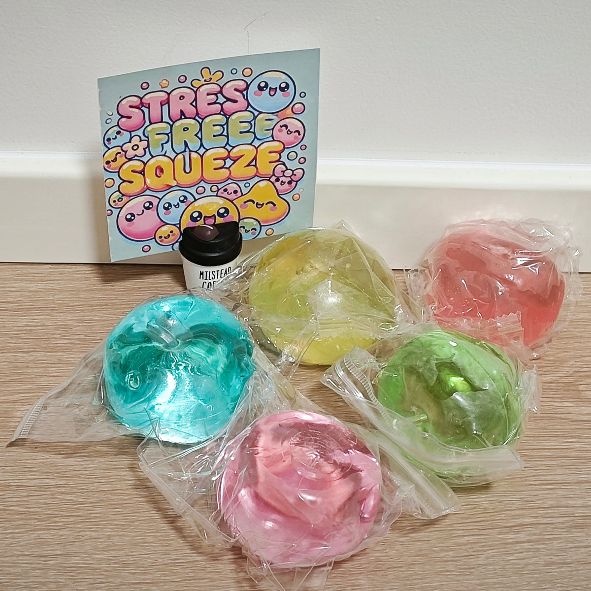 Five packaged pastel-colored stress balls arranged on a light wood floor in front of a white wall, with a card reading 'STRESS FREE SQUEEZE' and a small bottle behind them.