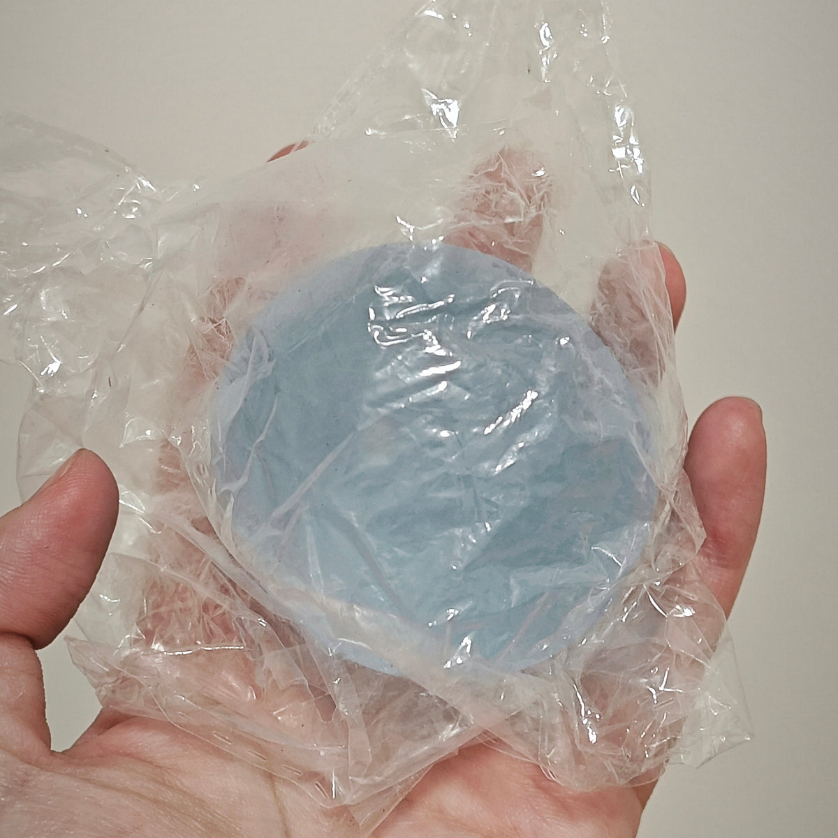 A hand holding a round, light blue item wrapped in clear plastic packaging against a plain light background.
