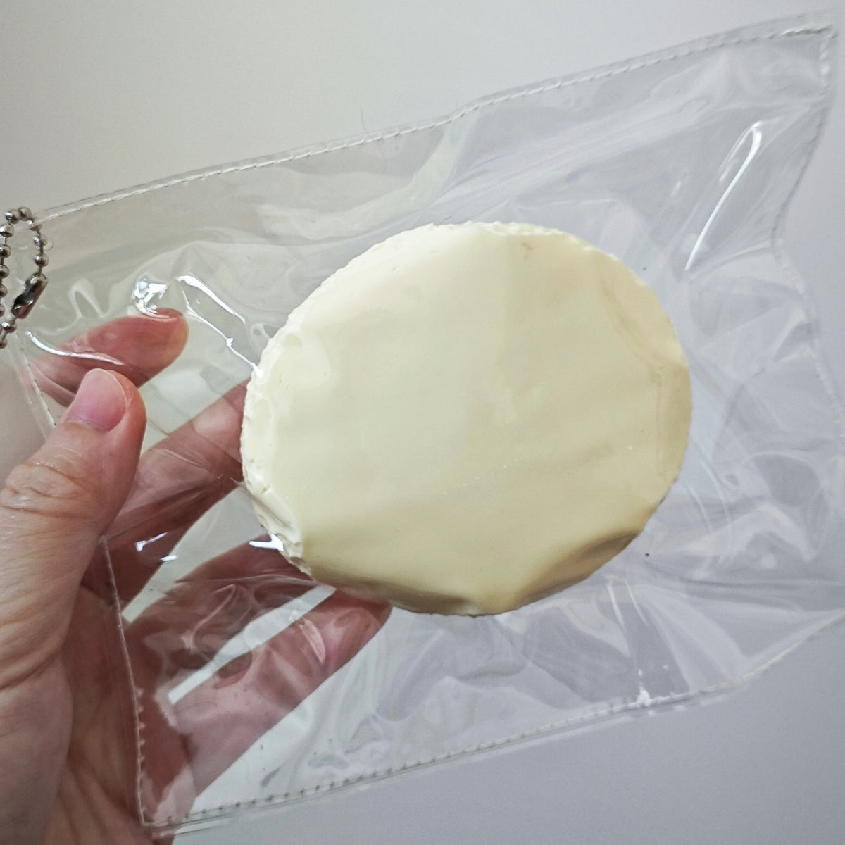 A hand holding a round, pale cream-colored item inside a clear plastic zip pouch against a plain light background.