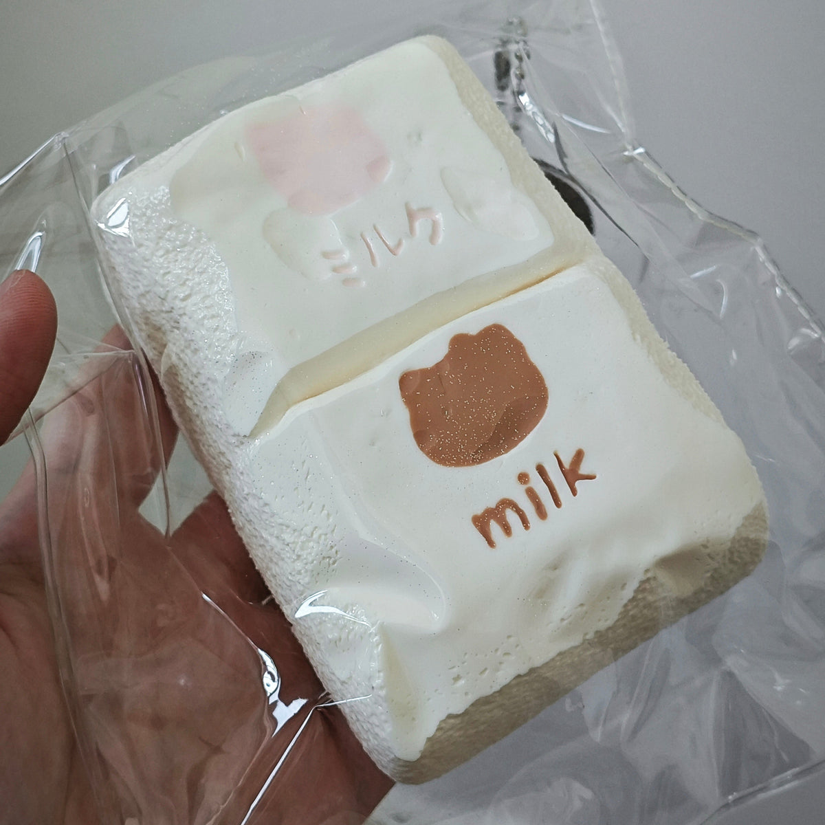 A hand holding a packaged squishy toy shaped like two stacked marshmallows, one white with a pink bear face and the other white with a brown bear face and the word 'milk' in brown.