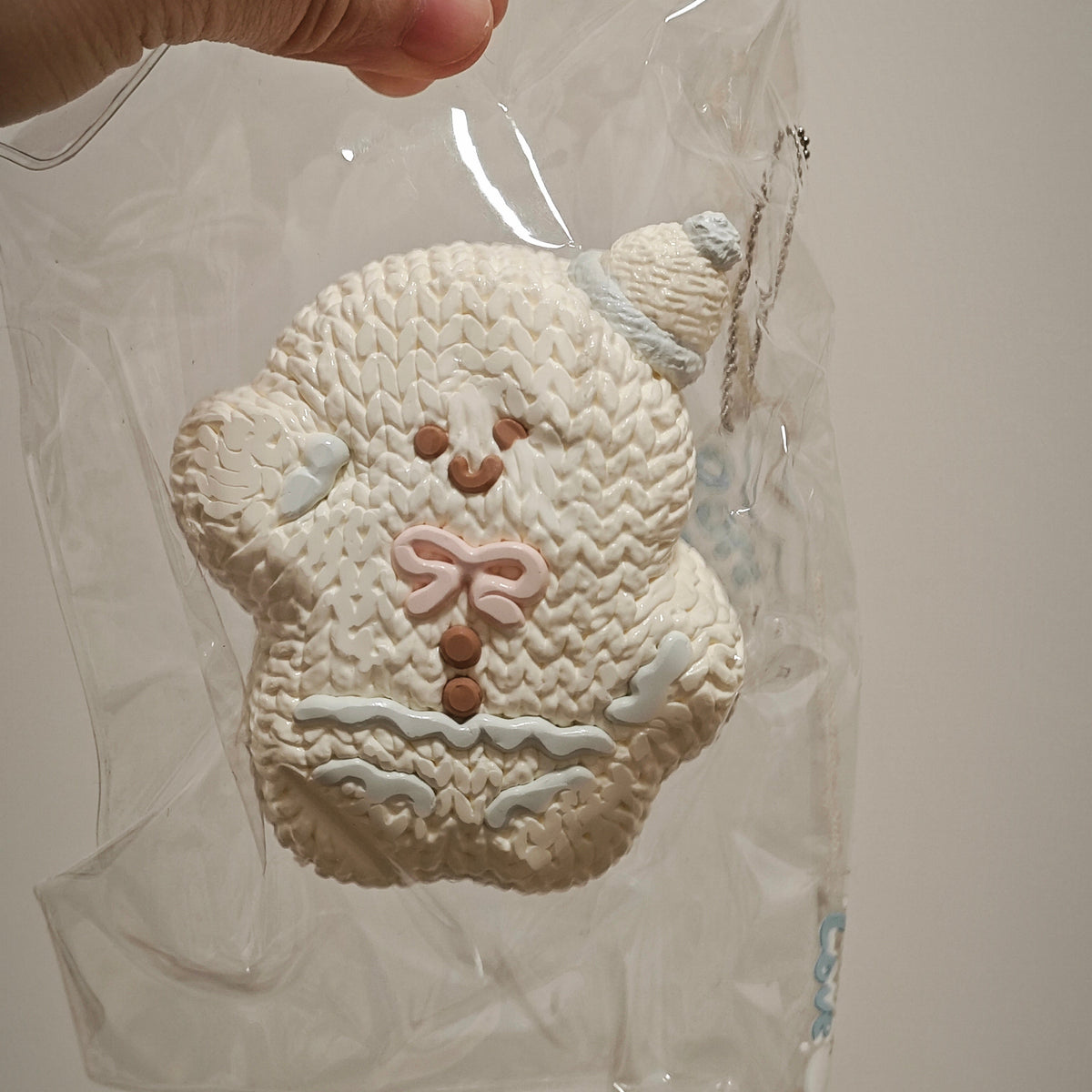 A hand holding a clear plastic bag containing a knitted snowman-shaped item, likely a cookie or decorative piece, against a plain light background.