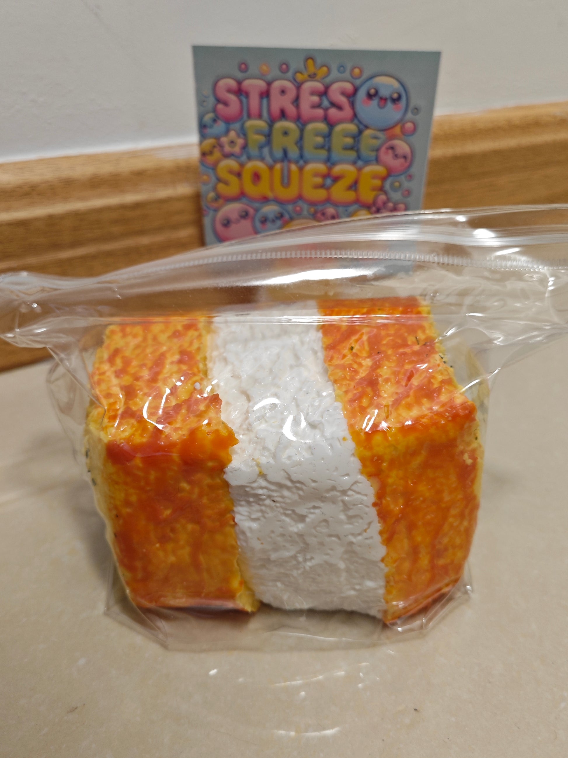 A packaged squishy toy shaped like a slice of bread or cake, with orange-yellow crust and a white center, sealed in a clear plastic bag. A card reading 'STRESS FREE SQUEEZE' is visible in the background.