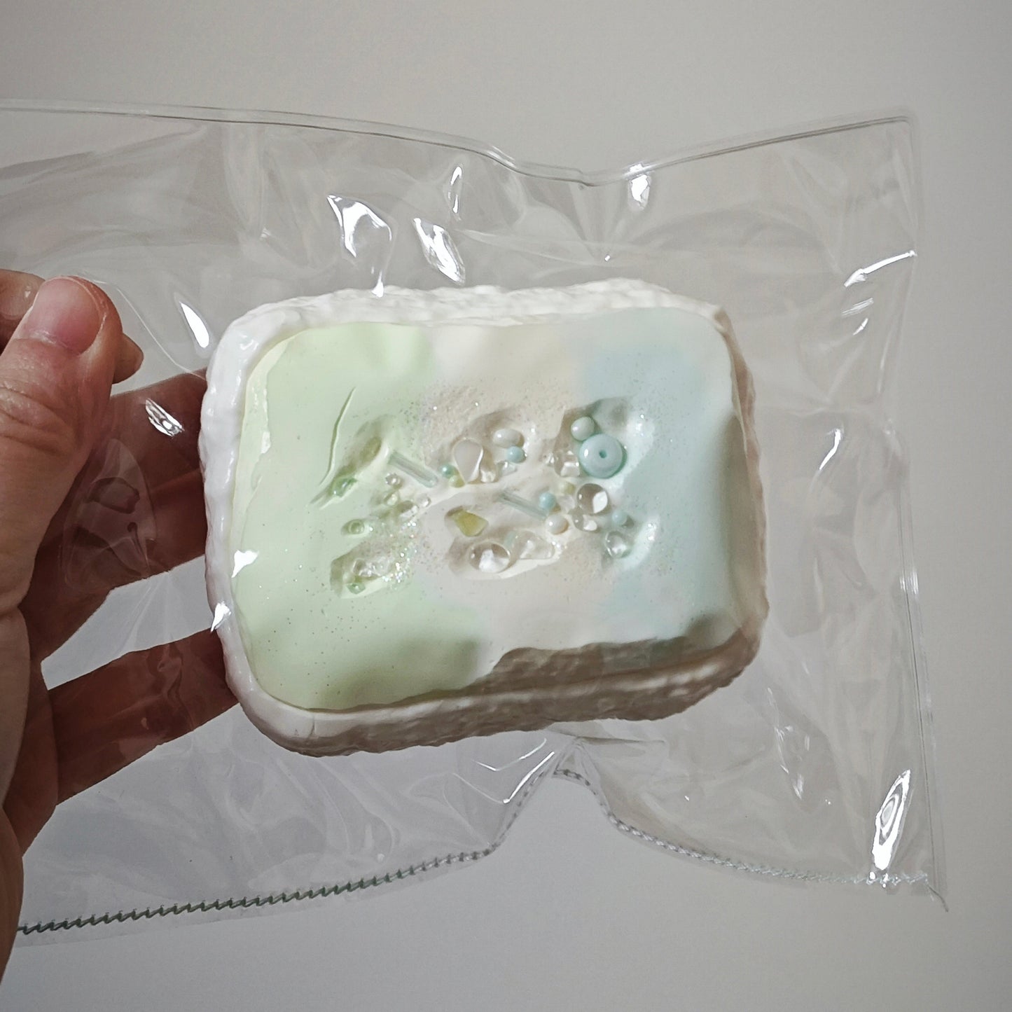 A hand holding a square, handmade soap in clear plastic packaging against a plain light gray background. The soap has a white base with a pale mint green and light blue floral pattern on top.