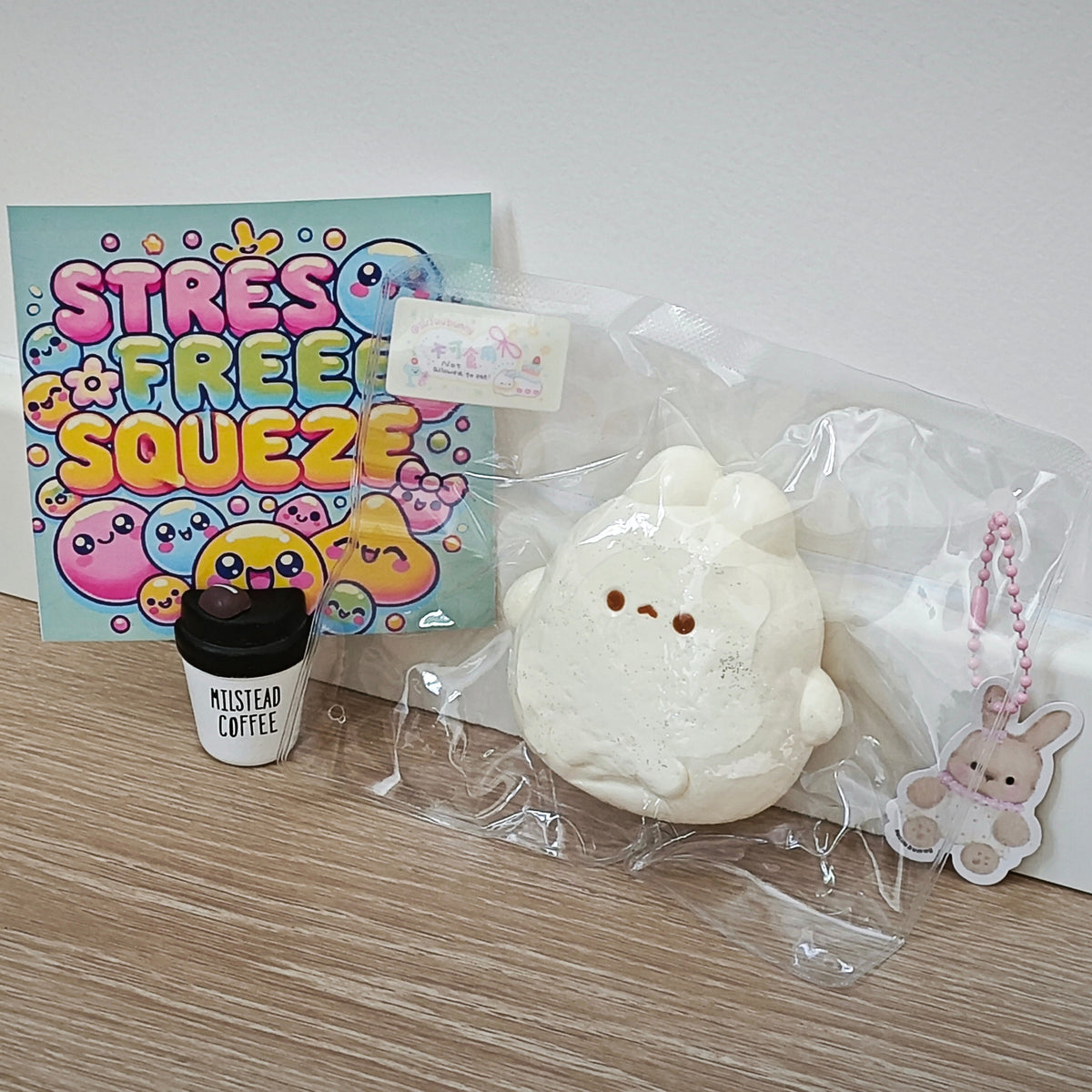 A white fluffy squishy toy in clear plastic packaging, displayed on a light wood floor next to a colorful 'STRESS FREE SQUEEZE' card, a small black-and-white cup labeled 'MISTYAR COFFEE', and a small bunny keychain.