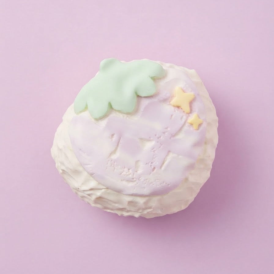 A pastel-colored strawberry-shaped item, possibly a bath bomb or decorative mold, featuring a light purple body, white ruffled edges, a mint green leaf on top, and two small yellow stars, set against a solid lavender background.