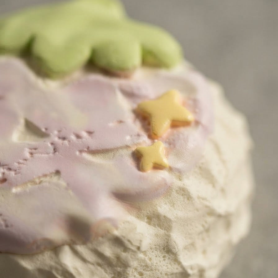 A close-up of a round, textured dessert or baked good topped with pale pink icing, a light green frosting section, and two small yellow star decorations, set against a soft gray background.