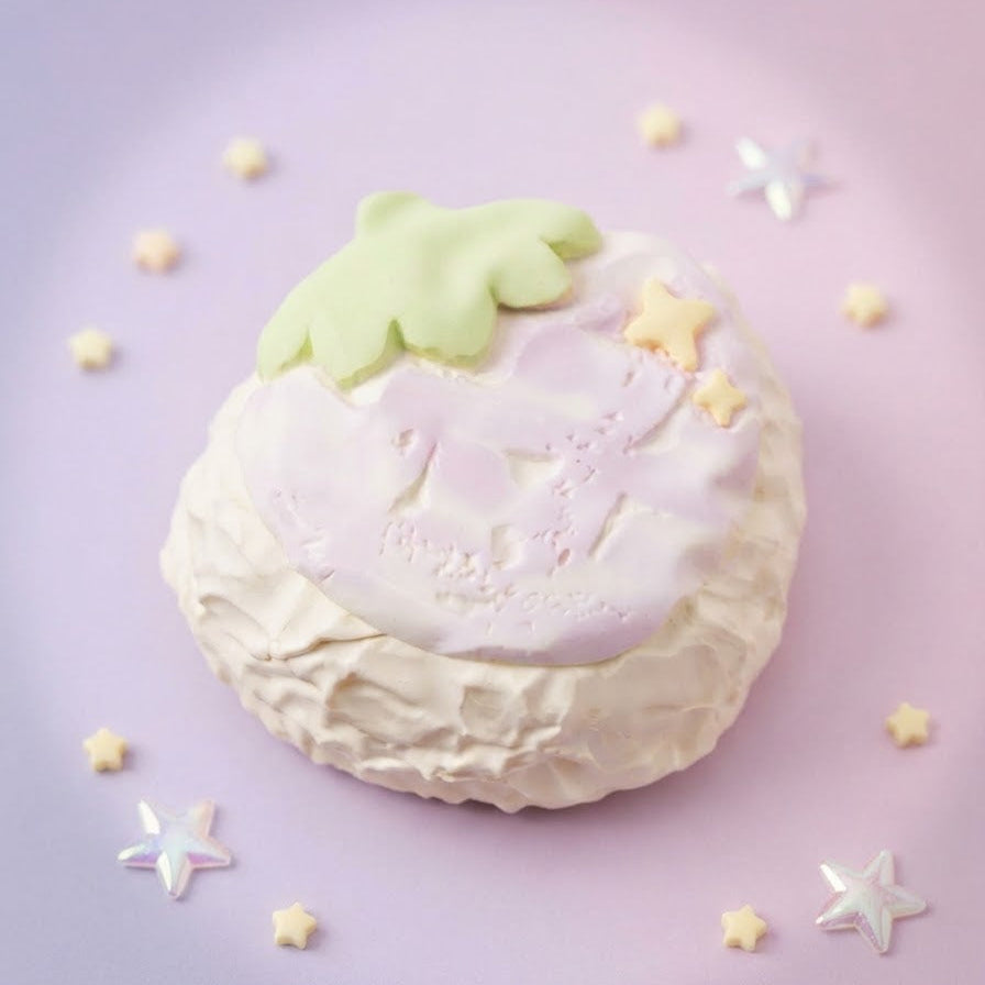 A pastel-colored strawberry-shaped cake or pastry on a soft pink background, decorated with a pale green leaf on top, a pink and white strawberry face, and small yellow star decorations.