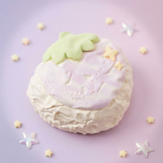 A pastel-colored strawberry-shaped cake or pastry on a soft pink background, decorated with a pale green leaf on top, a pink and white strawberry face, and small yellow star decorations.