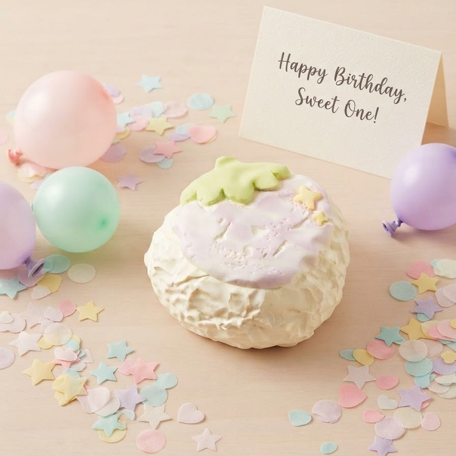 A pastel birthday cake decoration or foam topper shaped like a cupcake with a green leaf topper, placed on a light wood surface with colorful balloons, star and heart confetti, and a small card that reads 'Happy Birthday, Sweet One!'.