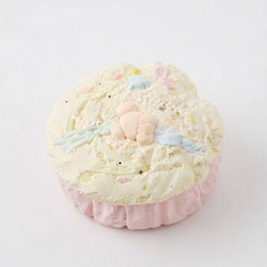 A pastel-colored bath bomb shaped like a cupcake, featuring a pale pink ruffled base, a textured off-white top with pastel speckles, and a small peach bunny decoration on top, set against a plain white background.