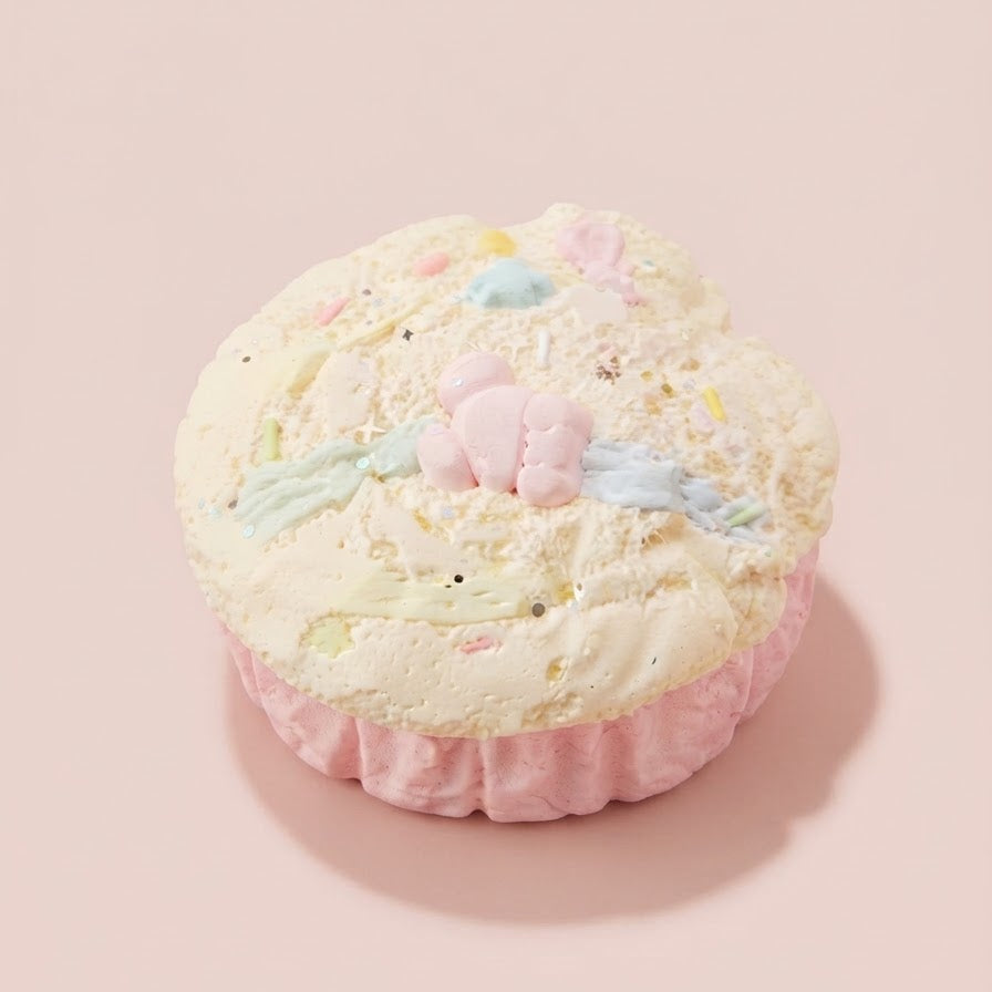A pastel pink cupcake-shaped product with white frosting-like top, decorated with small pink bunny figures, pastel blue and green accents, and scattered confetti, set against a solid pale pink background.