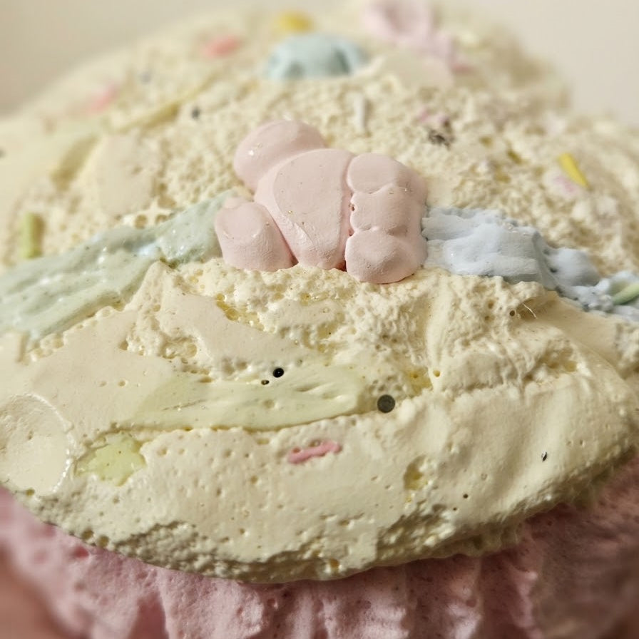 A close-up of a pastel-colored dessert, likely a meringue-based cake or cupcake, with pale yellow and pink frosting, sprinkles, and a small pink marshmallow bear decoration on top.