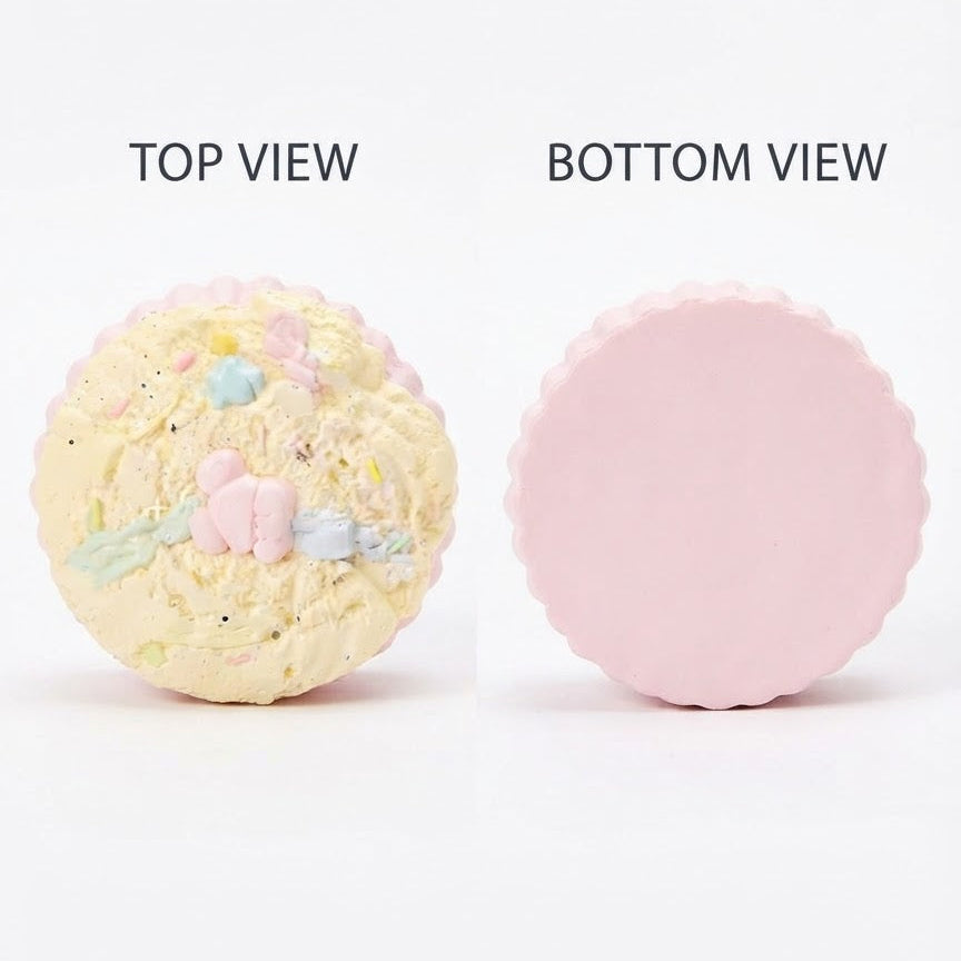 A round compact powder shown from two angles on a white background, labeled TOP VIEW and BOTTOM VIEW. The top view shows a pale yellow textured surface with pastel pink, blue, and green decorative details, while the bottom view shows a smooth light pink side.