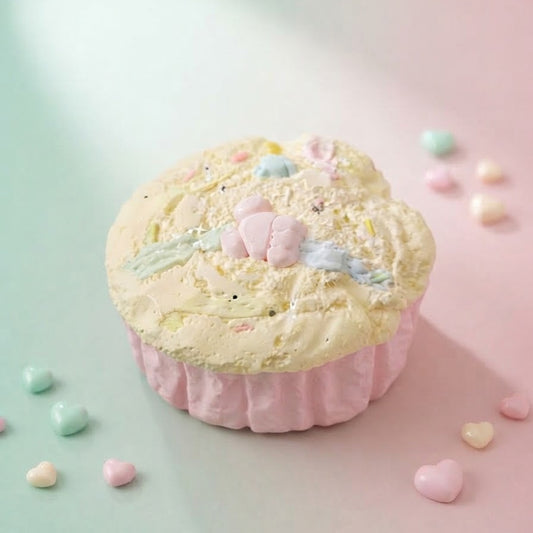 A pastel cupcake-shaped product with pale yellow frosting, pink wrapper, and small candy-like decorations, photographed on a soft mint and pink background with scattered heart-shaped candies.