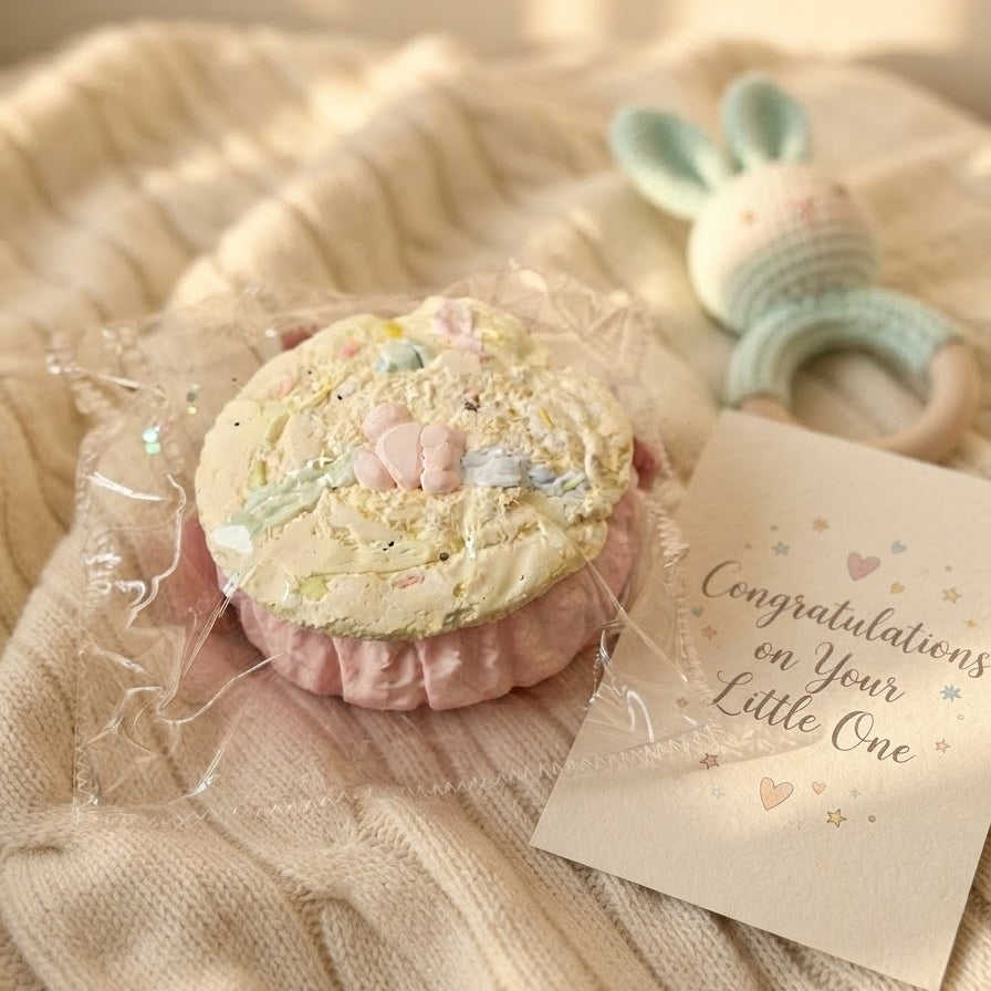 A round, pastel-colored baby keepsake or toy wrapped in clear cellophane, placed on a cream knit blanket next to a printed congratulations card and a small pastel rattle.