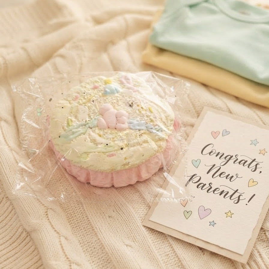 A pastel cupcake-shaped baby shower favor wrapped in clear cellophane, placed on a cream cable-knit blanket next to a 'Congrats, New Parents!' card and folded pastel garments.