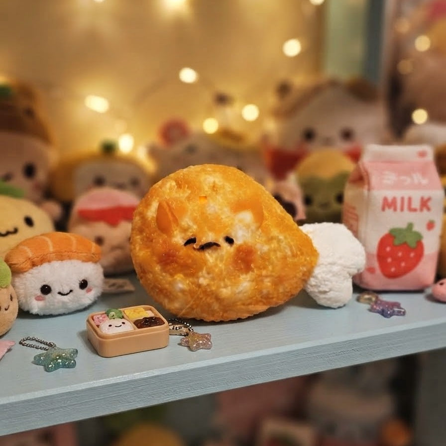 A cute, round, golden-brown plush toy shaped like a fried rice ball with a small face, displayed on a light blue shelf alongside other small plushies and accessories.