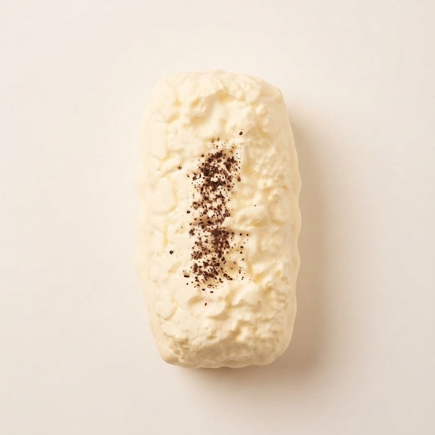 A single cream-colored textured bar, likely a soap or shampoo bar, topped with a vertical sprinkle of dark brown ground coffee, shown on a clean off-white background.