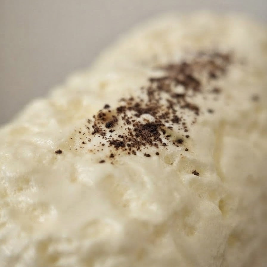 A close-up shot of a white, textured food item, possibly whipped cream or a creamy dessert, topped with a sprinkle of dark brown ground spice or cocoa powder.