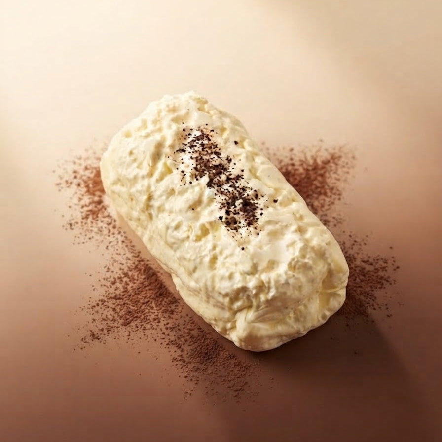 A cream-colored pastry or dessert roll topped with dark cocoa powder, presented on a warm brown surface with cocoa powder scattered around it.