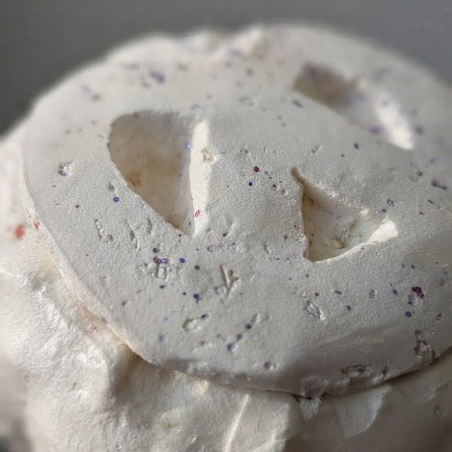 A close-up of a white sculptural object with a rough, textured surface and indented triangular and oval shapes, speckled with tiny multicolored flecks, against a neutral gray background.