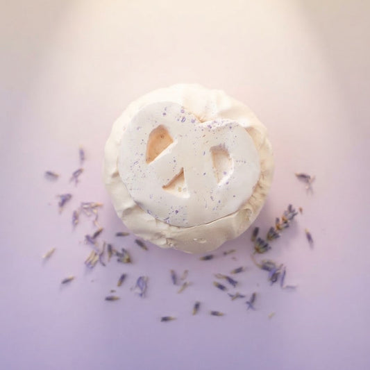 A round white bath bomb with a cutout peace sign, decorated with purple glitter, resting on a lavender surface with scattered dried lavender sprigs.