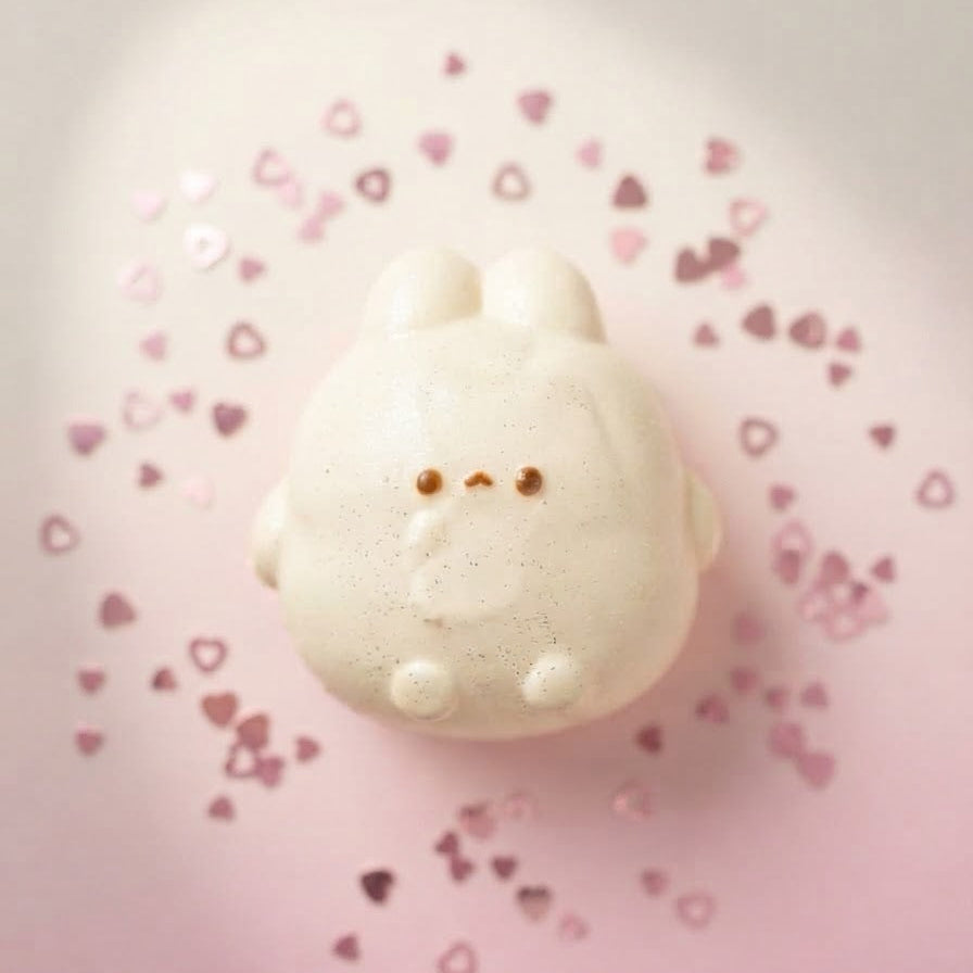A small white bunny-shaped decorative object with tiny pink heart-shaped confetti scattered around it on a soft pink background.