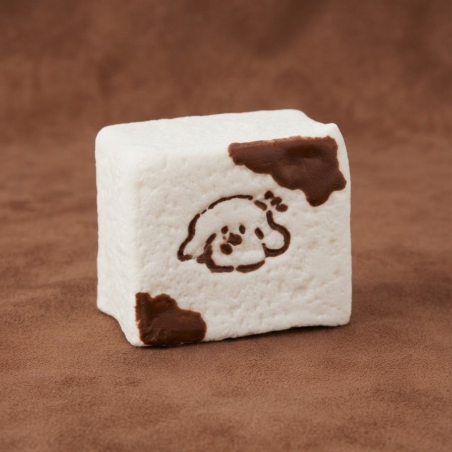 A square white marshmallow or foam block with brown cow-print spots and a simple brown dog face illustration, photographed on a brown textured background.
