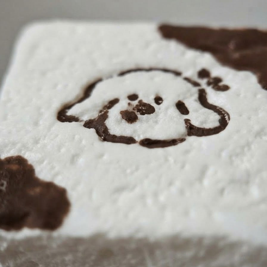 A close-up of a white square product, likely a soap, candle, or decorative block, featuring a dark brown hand-drawn dog face and paw print design.