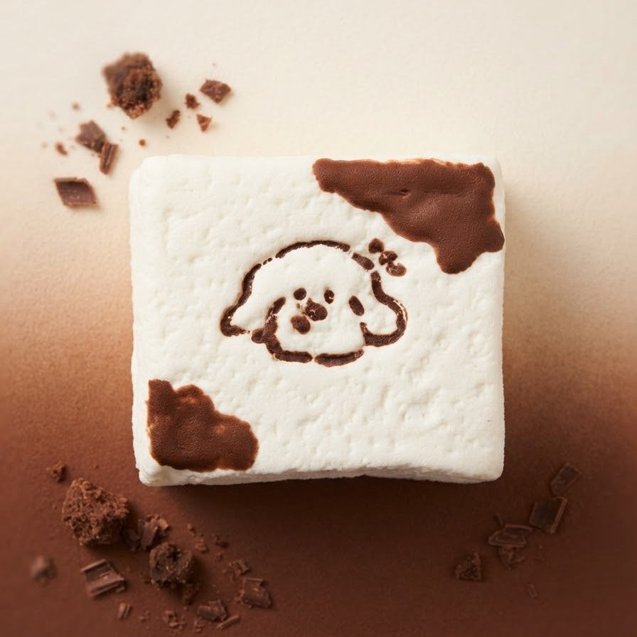 A square white marshmallow treat with chocolate-brown chocolate splatters and a small dog face design in the center, photographed on a warm beige-to-brown gradient background with scattered chocolate crumbs.