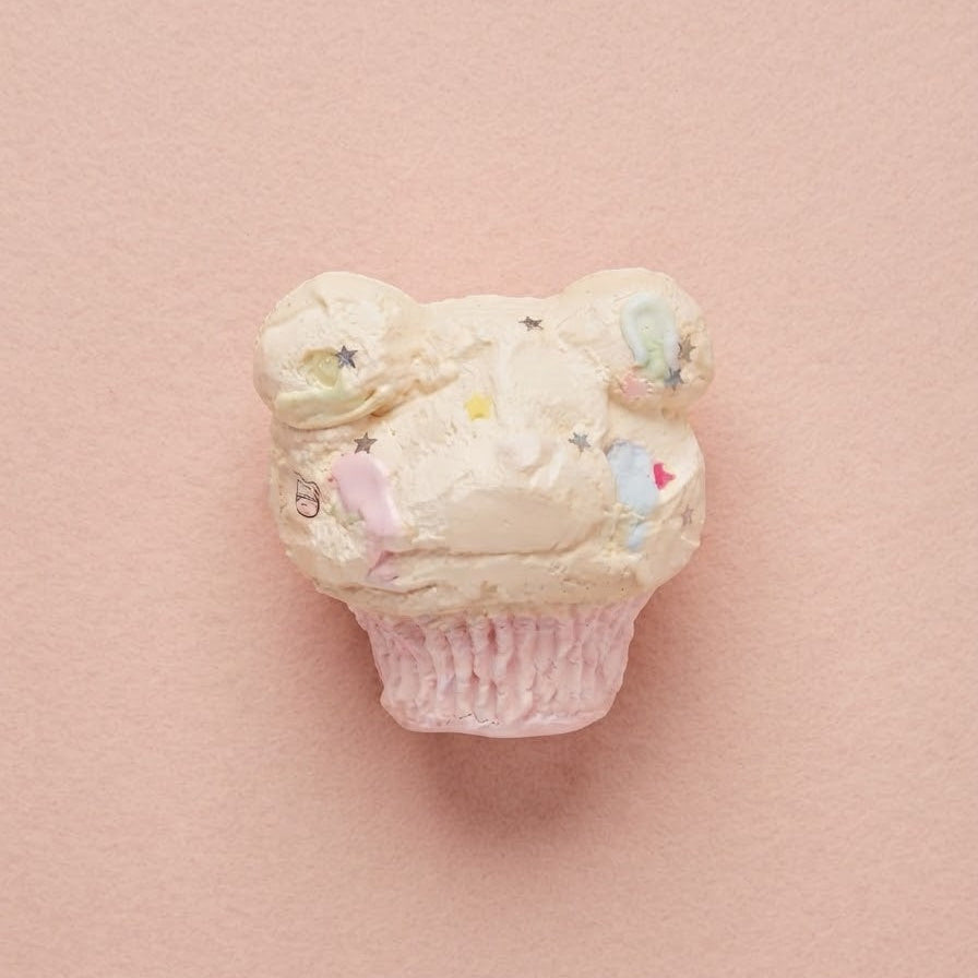 A small cupcake-shaped decorative object with pale pink frosting, tiny pastel accents, and a pink liner base, centered on a soft pink background.