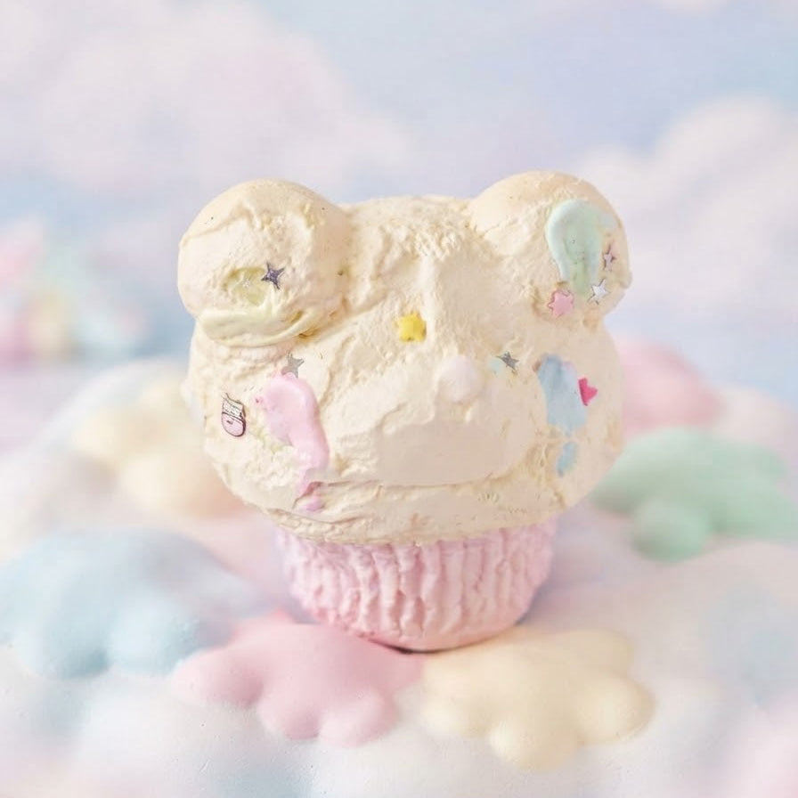 A pastel cupcake-shaped product with cream-colored frosting topped with bear ears and small multicolored star and heart sprinkles, set on a soft cloud-like surface against a dreamy pastel background.