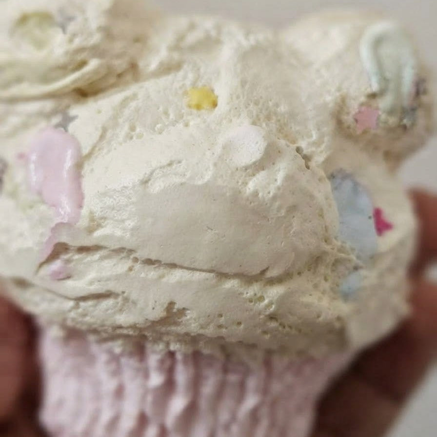 A close-up of a cupcake topped with thick, textured off-white frosting and small pastel decorative accents, held in a hand against a plain light background.