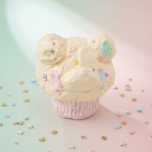 A pastel cupcake-shaped product with cream-colored frosting topped with bear ears and small multicolored star and heart decorations, sitting on a pink cupcake liner against a soft mint and pink background with scattered star confetti.