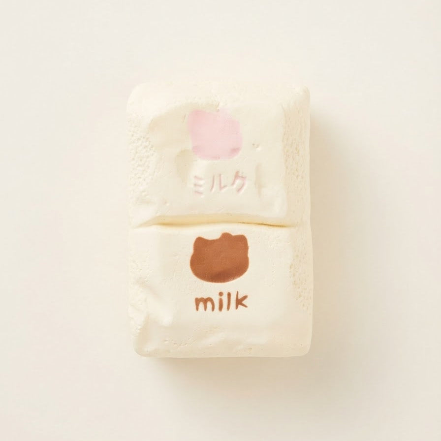 Two small cream-colored packaged items stacked vertically on a plain off-white background. The top package has a light pink shape and Japanese text, while the bottom package has a brown shape and the word "milk" in English.