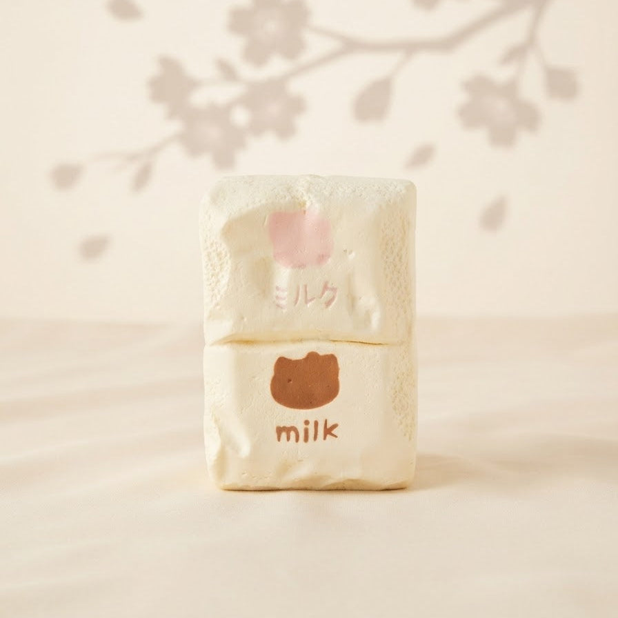 A small stacked set of two cream-colored textured blocks, one with a pink cherry blossom graphic and Japanese text, and the other with a brown bear face graphic and the word 'milk' in brown. The set is displayed on a light beige surface with a soft shadow of cherry blossoms cast in the background.