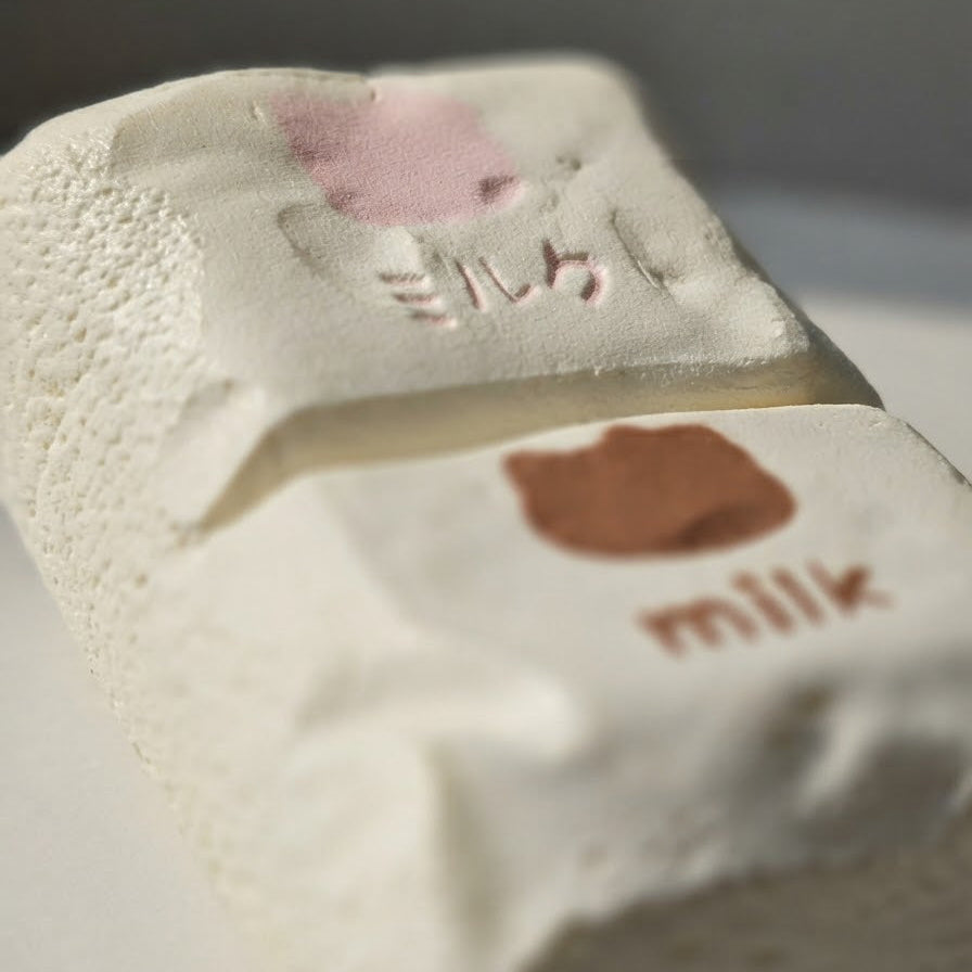 A close-up of a textured white block with embossed pink and brown shapes and the word 'milk' printed on it, photographed against a soft gray background.