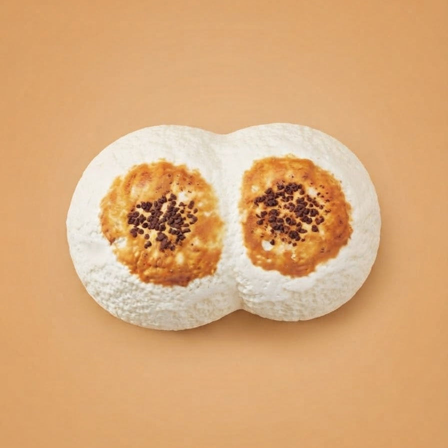 Two white, dough-like novelty objects shaped like a double bubble with toasted golden-brown circular spots and dark sprinkles on top, set against a solid warm peach background.
